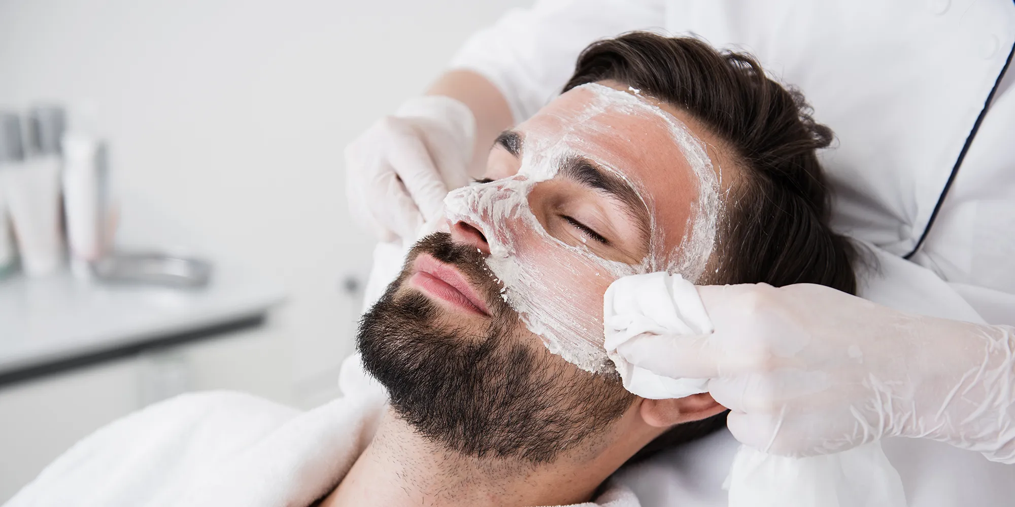 Close-up van een man met gesloten ogen die een gezichtsbehandeling krijgt, waarbij een specialist met handschoenen een witte crème op zijn gezicht aanbrengt.