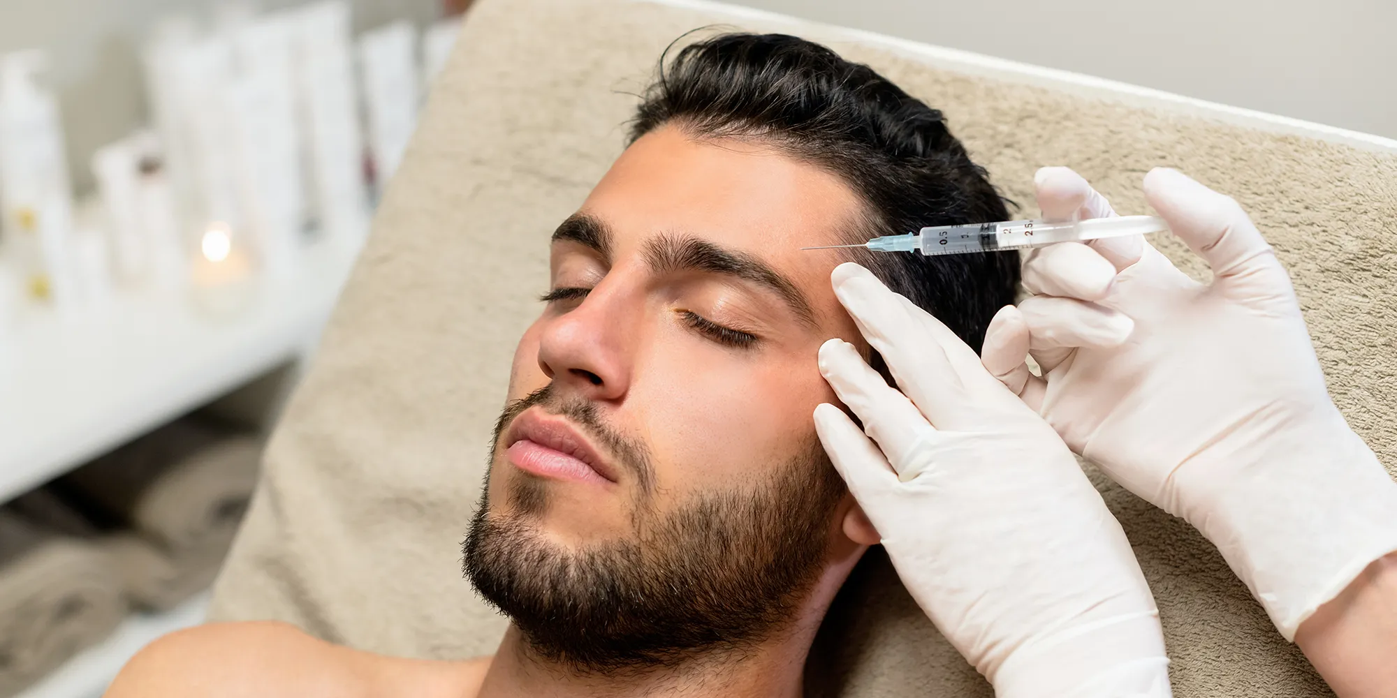 Close-up van een man met gesloten ogen die een cosmetische injectie in het voorhoofd krijgt toegediend door een specialist met handschoenen.